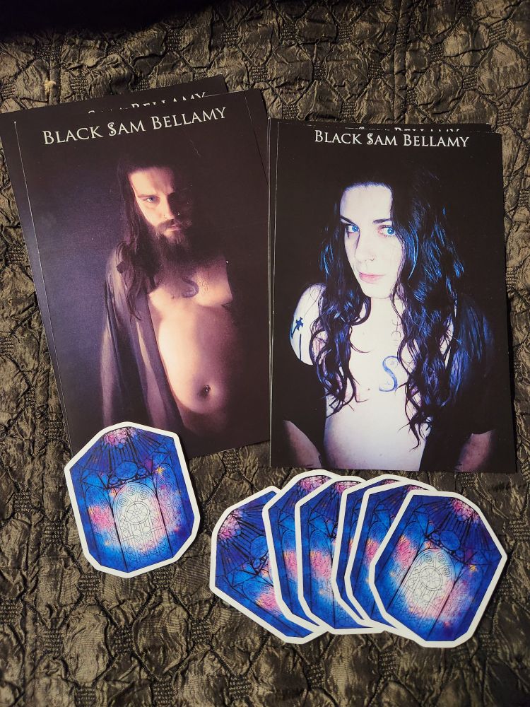 Left is a masculine print of Sam looking intensely at the camera in a sheer robe. Right is a feminine print of Sam, blue eyes vibrant and hair stark cascading over chest. Underneath the prints are stickers of stained glass lanterns filled with galaxies.