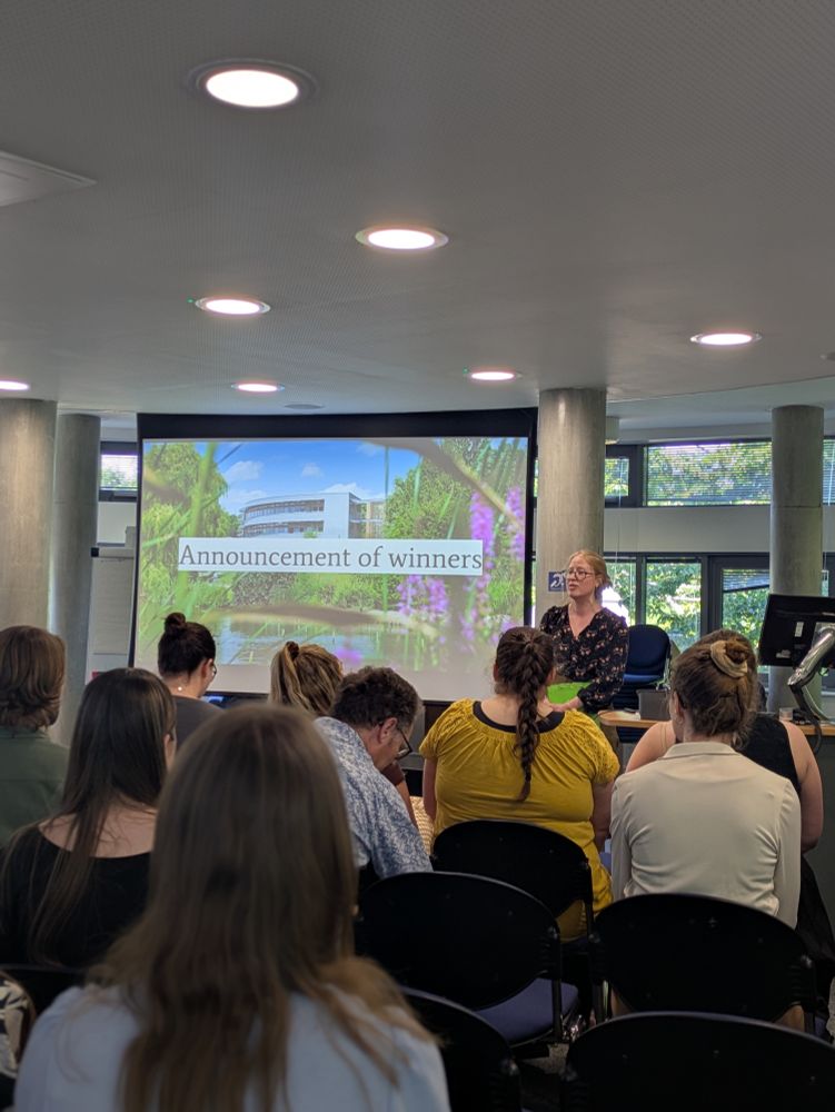 Photo taken from the back of the room, we can see the seated audience and Karen Baker announcing the winners next to a screen that says 'Announcement of winners'