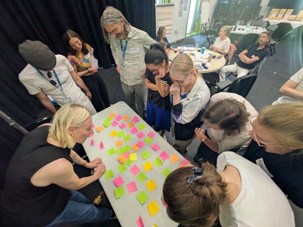 A group of workshop participants standing over a table looking at post-it notes and thinking
