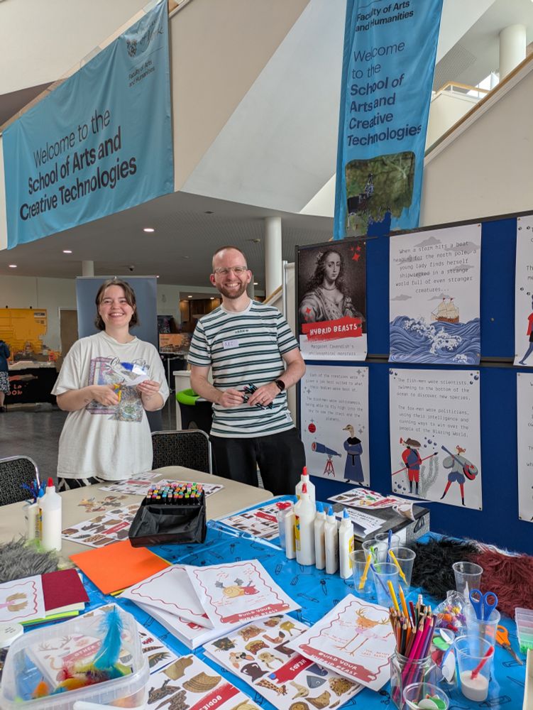 Sophie White and colleague at their exhibition stand. There's lots of cutouts, as well as glitter, pencils, feathers, and posters in the background. 