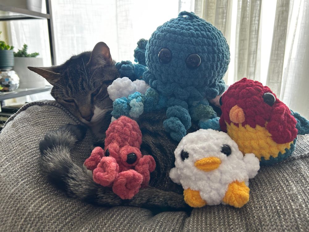 A picture of cam (a gray tabby cat) sleepily curled up with a pile of crochet stuffed animals. 