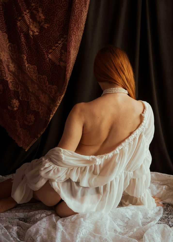 Photo d'une femme de dos dans un décor en studio fait de rideaux et de voilages. Elle porte une blouse qui tombe sur son épaule 