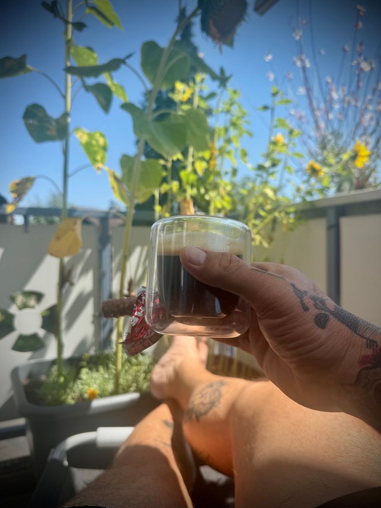 Ein Mensch sitzt entspannt in der Sonne auf einem Balkon, die Beine ausgestreckt, und hält eine kleine, transparente Tasse mit schwarzem Kaffee und einem Schoggistängeli in der tätowierten Hand. Im Hintergrund stehen hohe Sonnenblumen in Blumentöpfen, ihre Blätter und Blüten leuchten grün und gelb im warmen Tageslicht. Die Atmosphäre wirkt ruhig, sommerlich und entspannt.
