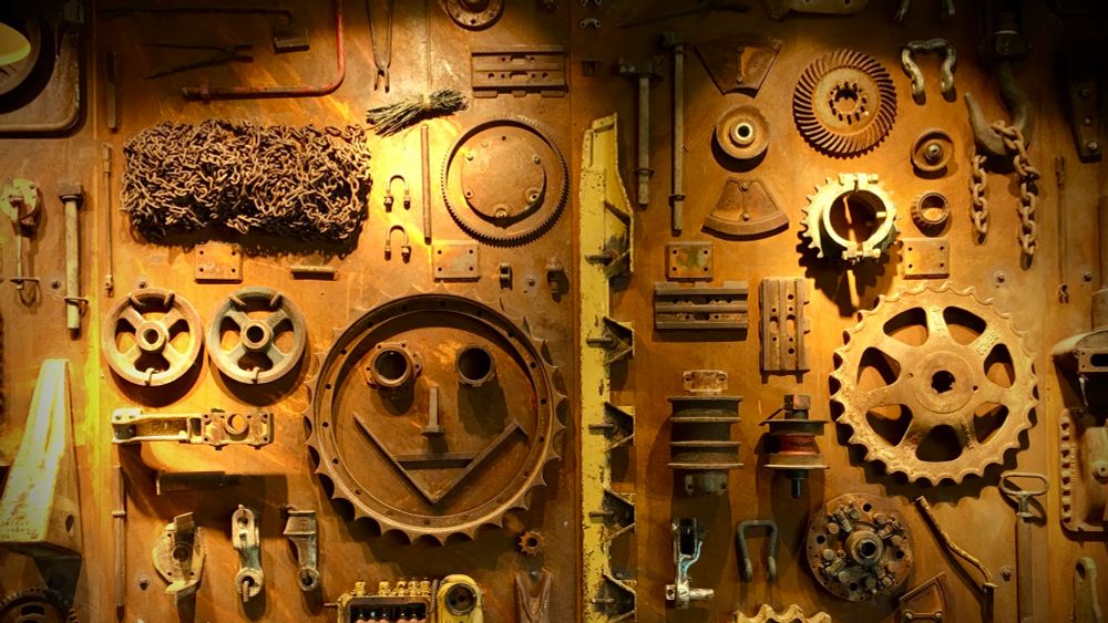 Eine Wand voller alter, rostiger Maschinenteile und Zahnräder ist kunstvoll arrangiert. Einige Teile bilden das Bild eines lächelnden Gesichts aus einem großen Rad und Schrauben als Augen, andere Elemente sind chaotisch, aber ästhetisch angeordnet, darunter Ketten, Zahnräder und Werkzeuge. Das Licht betont die Metallstrukturen und den industriellen Charme der Szene.