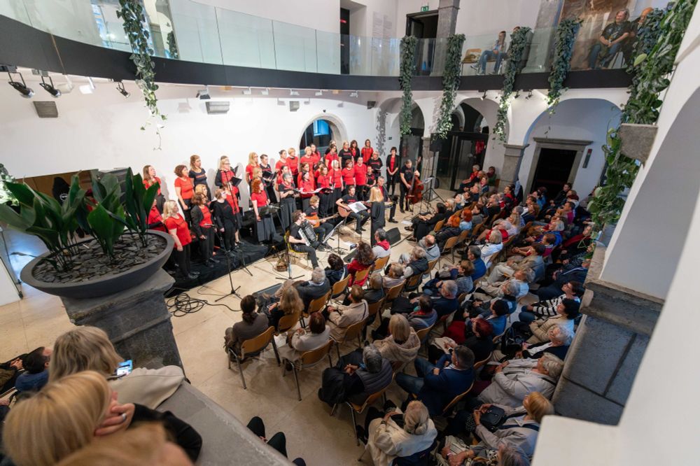 Zbor z več kot 30 pevkami poje na odru, pred njimi so štirje glasbeniki z instrumenti (harmonika, kitara, bobni in kontrabas), dirigentka vodi zbor s hrbtom proti občinstvu, ki sedi na stolih in stoji na balkonu svečane dvorane.