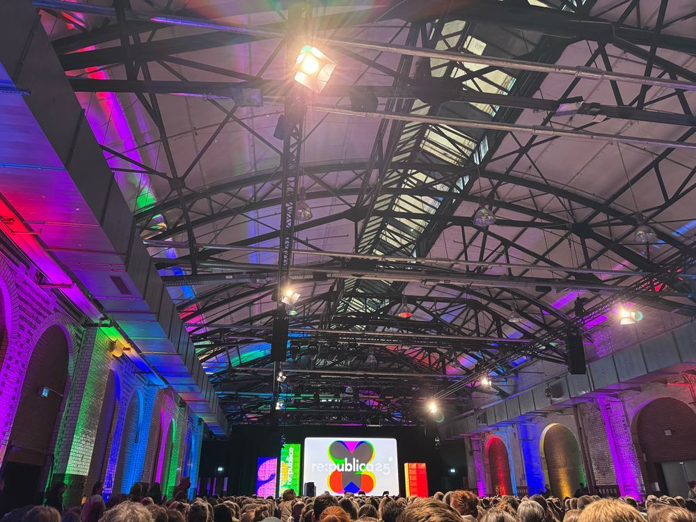 Große Veranstaltungshalle mit industrieller Architektur und gewölbtem Dach, gefüllt mit vielen Menschen. Die Wände sind mit bunten Lichtern in Regenbogenfarben beleuchtet. Im Hintergrund befindet sich eine große Leinwand mit dem Logo und Text „re:publica 25“.