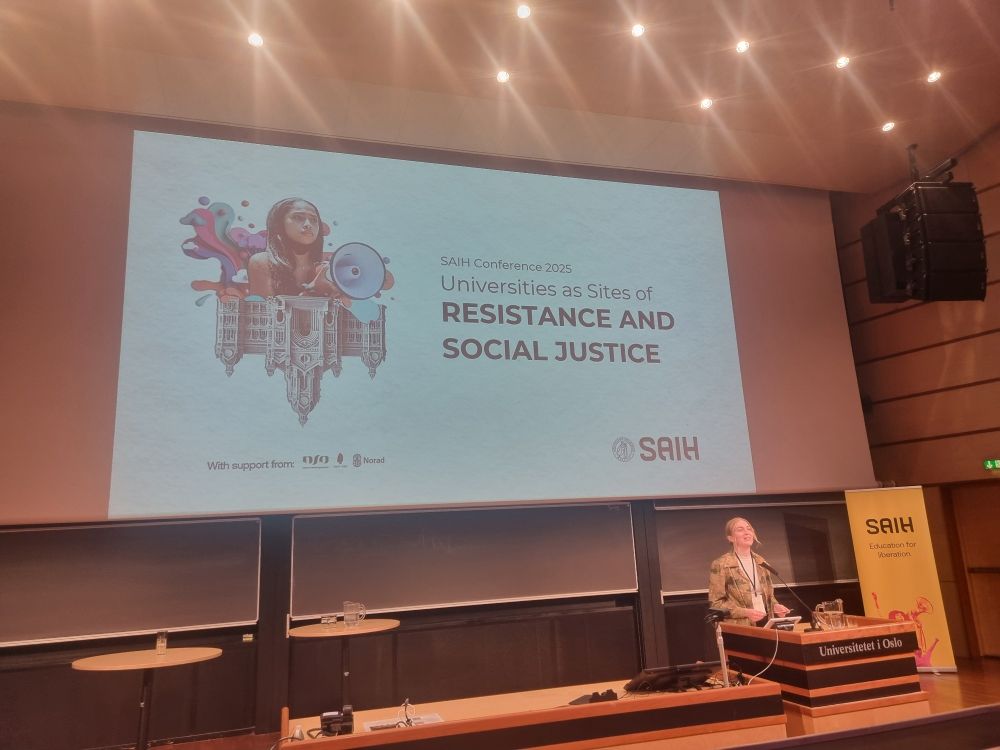 Selma Bratberg, President of the organisation SAIH, speaking in front of a large screen with the text "SAIH Conference 2025: Universities as sites of resistance and social justice" and an illustration with an image of a dark-haired woman with a megafone over an upside-down, stately building. 