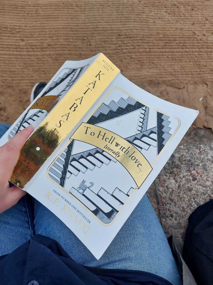 A proof copy of Katabasis by R. F. Kuang held open on my lap with the pages towards my leg, as I'm sat on a wall on the beach