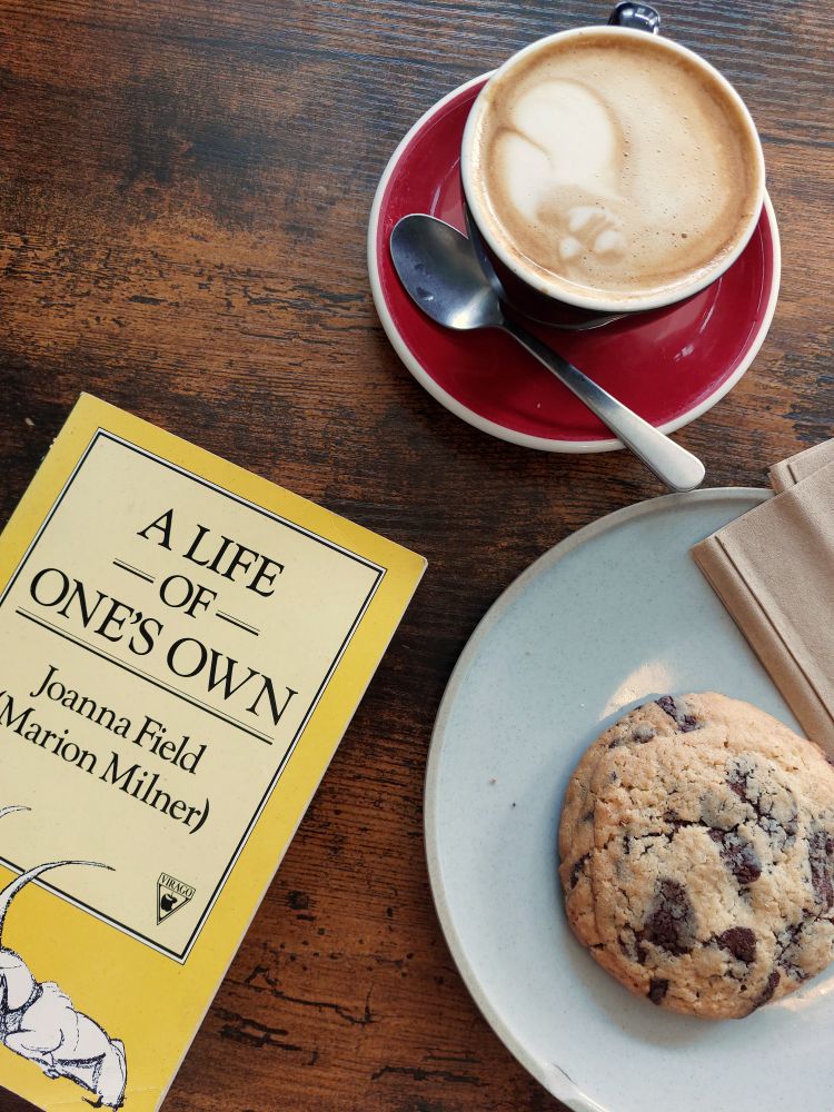 The Virago edition of A Life Of One's Own by Marion Milner (published under the pseudonym Joanna Field) lies on a wooden table. At the top of the table is a flat white coffee on a red saucer and underneath that there is a very large chocolate chip cookie on a plate.