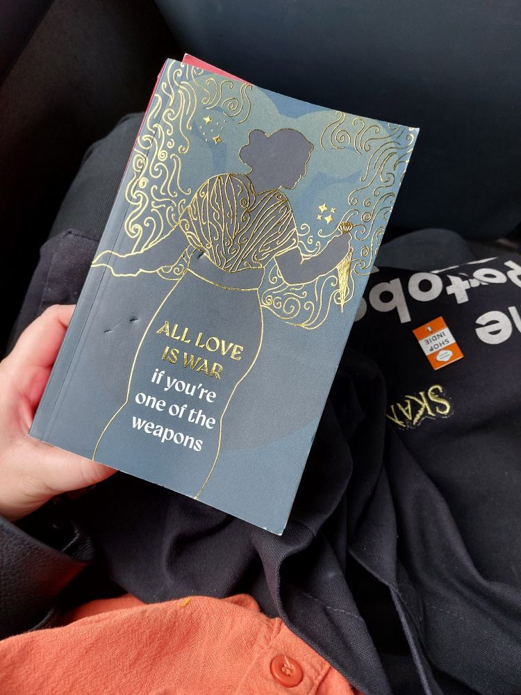 A proof copy of No Friend To This House by Natalie Haynes held by my hand over my lap on the bus, with my tote bag decorated with pins and badges in the background