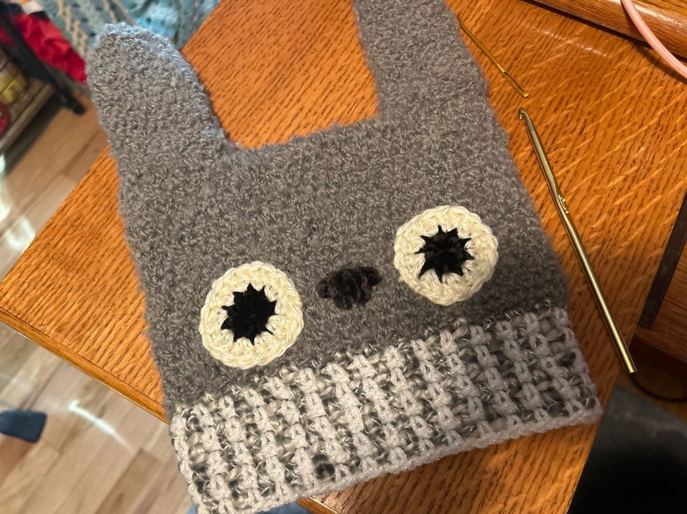 Finished crochet hat that looks like Totoro on a desk: grey rabbit with round eyes, a black velvet nose, and a grey and white band. Crochet hook and needle on its right 