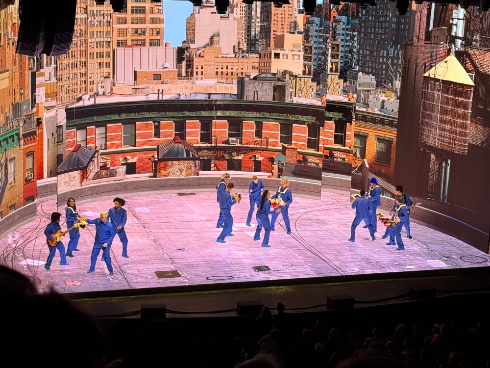 Photo of a David Byrne concert. The stage and backdrop are an LED of an NYC rooftop. The band all has wireless instruments and blue jumpsuits.