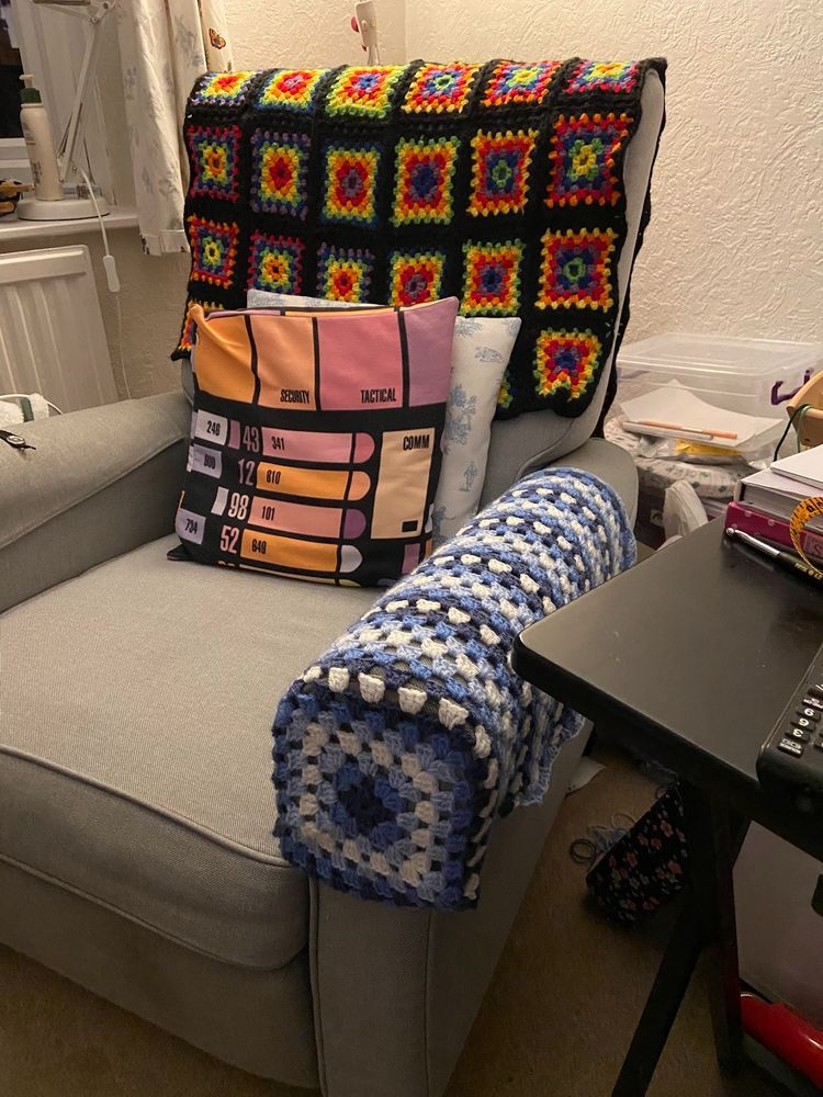 A recliner chair with a multi-rainbow and black granny square blanket on the back, Star Trek and Star Wars themed cushions, and a granny square armrest cover in three shades of blue plus white.