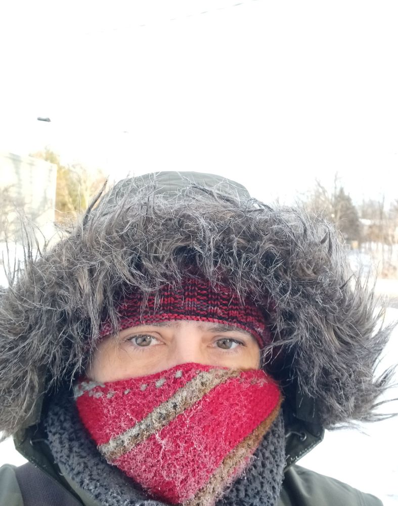 Headshot if a person bundled in a parka with faux fur trim over a touque and a frosted scarf