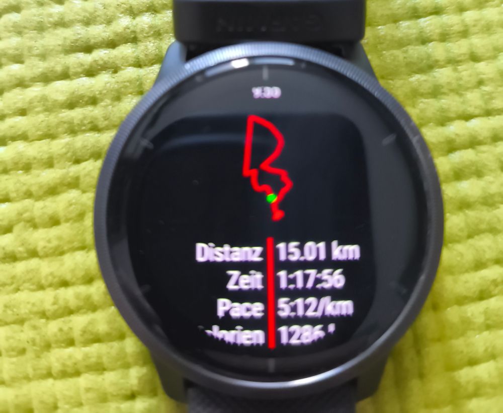 Schwarze Uhr auf grüner Matte. 
Die Anzeige:
Distanz 15.01 km
Zeit 1:17:56
Pace 5:12/km