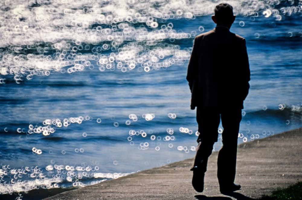 Taken autumn 1990. A lone figure, shot from behind, on the riverside walk by the River Tay, near Broughty Ferry. Landscape format; the figure is to the right of the image, in near silhouette, wearing a suit, hands in trouser pockets. Walking along a concrete path that crosses the frame diagonally, roughlt from bottom left to 1/3 up the right side. The the figure's left is river, blue, with out of focus highlights. The highlights are shown as small, white rings or 'doughnuts', a characteristic of the Tamron lens used to take the photo: a 500mm 'catadioptric' or mirror lens, light and compact, its design caused the highlights to take on this ship - you either like or don't like it ! Camera was an Olympus OM2SP, film was Fuji Velvia - then quite new.