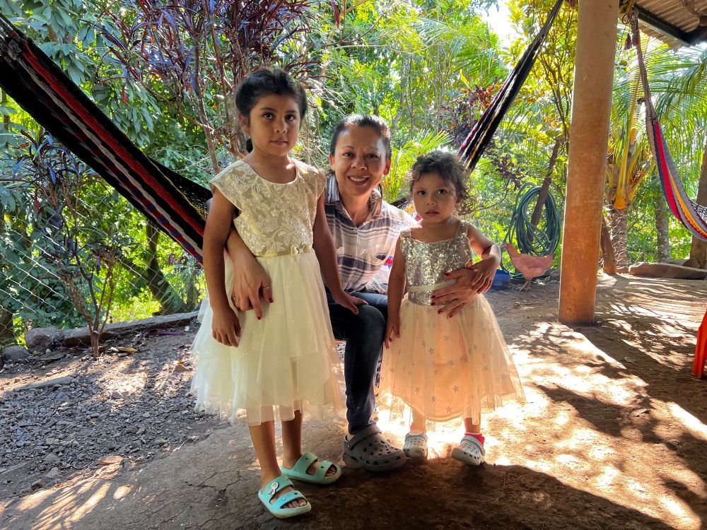 A warm family portrait of a woman smiling while kneeling beside two young girls wearing elegant white dresses. They are in an outdoor area surrounded by lush greenery, hammocks, and a cozy, natural atmosphere. A golden hen poses in the background. 