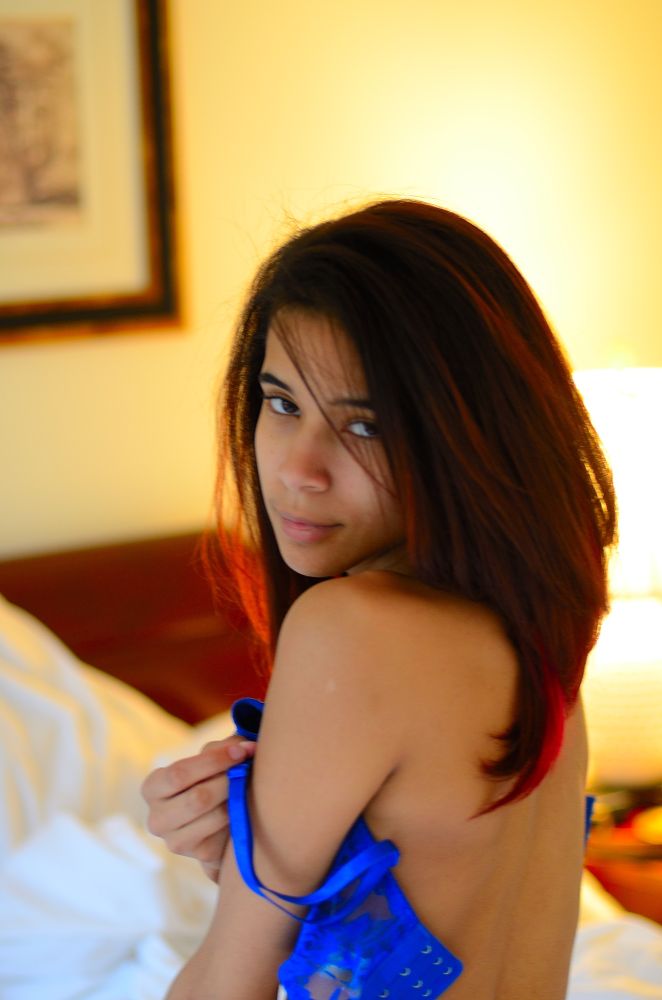 A young woman who is hispanic, slips off her blue bra while in bed, glancing at the camera