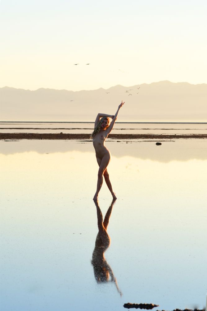 A nude blonde stands in the shallows of the Great Salt Lake in Utah, with her impade reflected in the water.