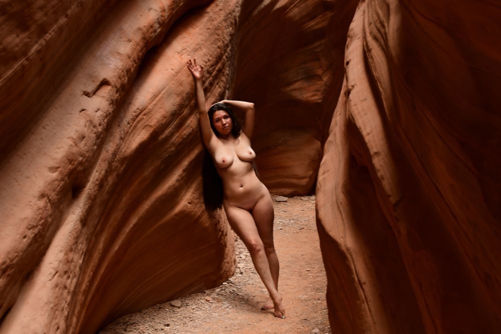 A nude busty latina with long black hair poses in a slot canyon.