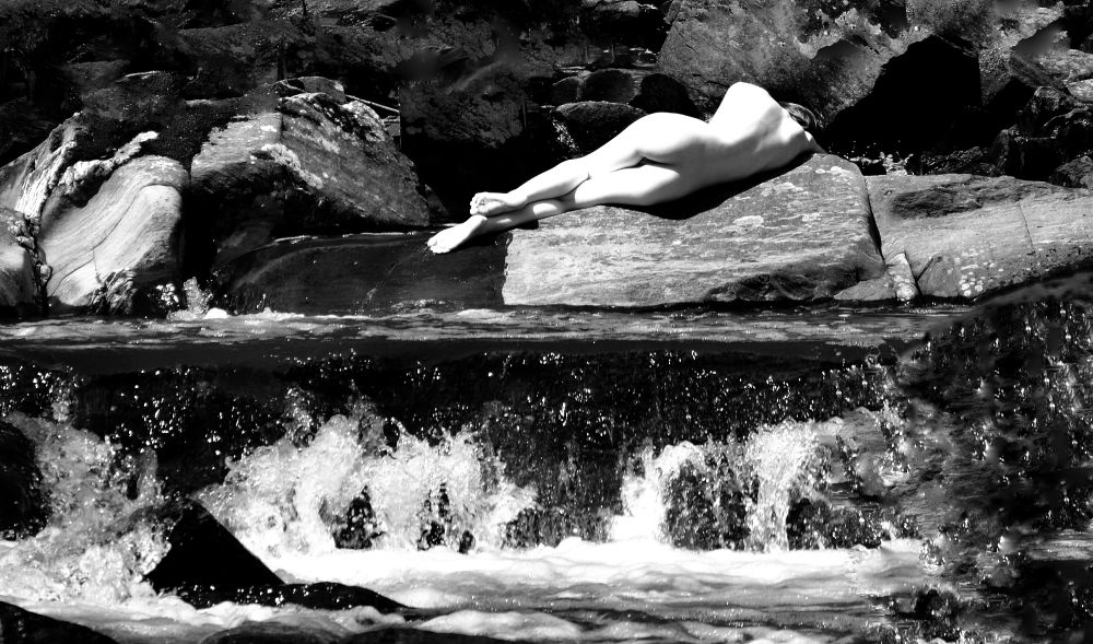 A nude woman poses on top of a rock in the middle of a stream with her back to the camera, in this black and white contrasting photo.