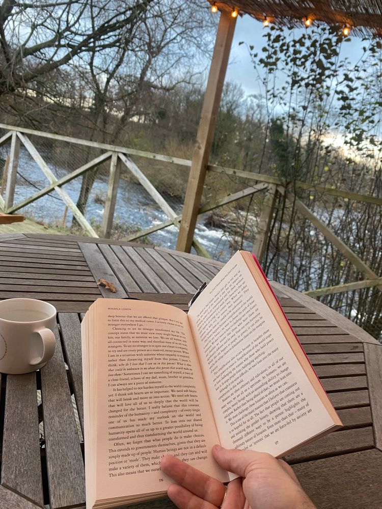 Man holding open a copy of ‘It’s Not That Radical’ by Mikaela Loach on an outdoor coffee table overlooking a river