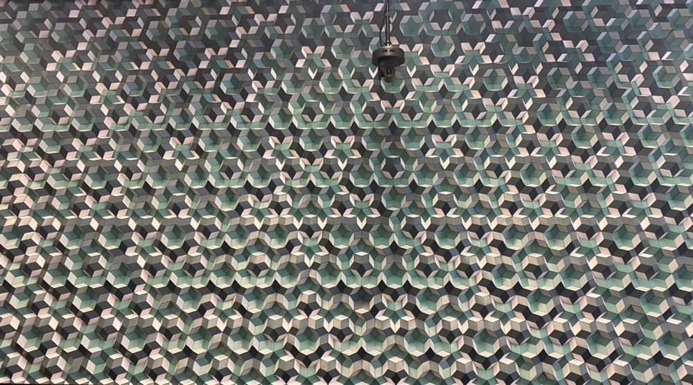 A view of the wall (only part of it) but zoomed out to show its aperiodic structure. It looks like a 3D variant of the aperiodic Penrose tiling, which is built out of a couple of kinds rhombus, which are then arranged into star shaped tessellates. The colors are white, a cool blue green and charcoal gray.
