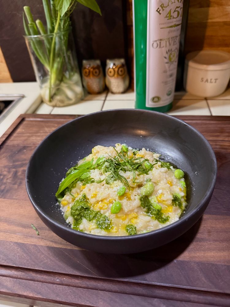 A black bowl on a dark wooden cutting board of sweet corn risotto with edamame, a little sharp and smoked provolone, pecorino romano, fresh pesto and lemon basil garnish. 