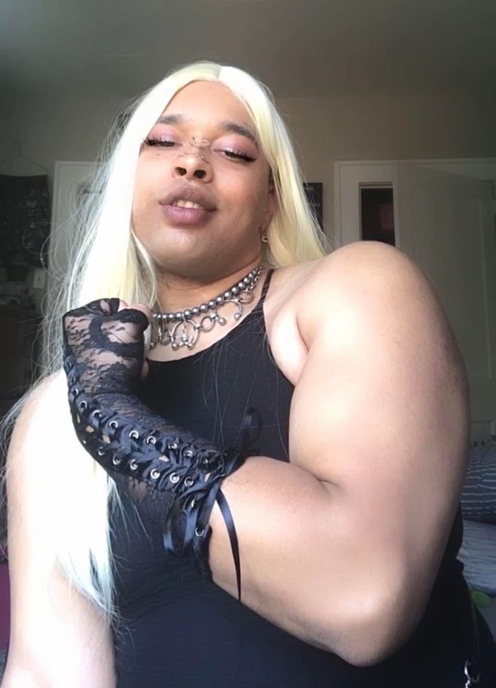 Buff muscle mommy trans girl in a blonde wig with a black dress flexing her bicep and smirking at the camera 