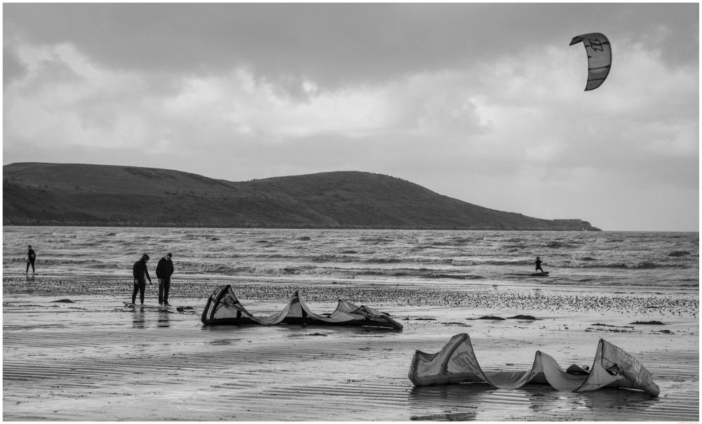 A black and white winter beach scene on a windy Bristol Channel. A bystander shows interest as a surfer explains the basics to him - middle left of the frame.

There is a para-surfer scudding across choppy water behind and to the middle right of the guys on the shore.

In the background can be seen the Brean Down Peninsula and on the end, Brean Down Fort.

The sky is cloudy though there is some brighter light breaking through.