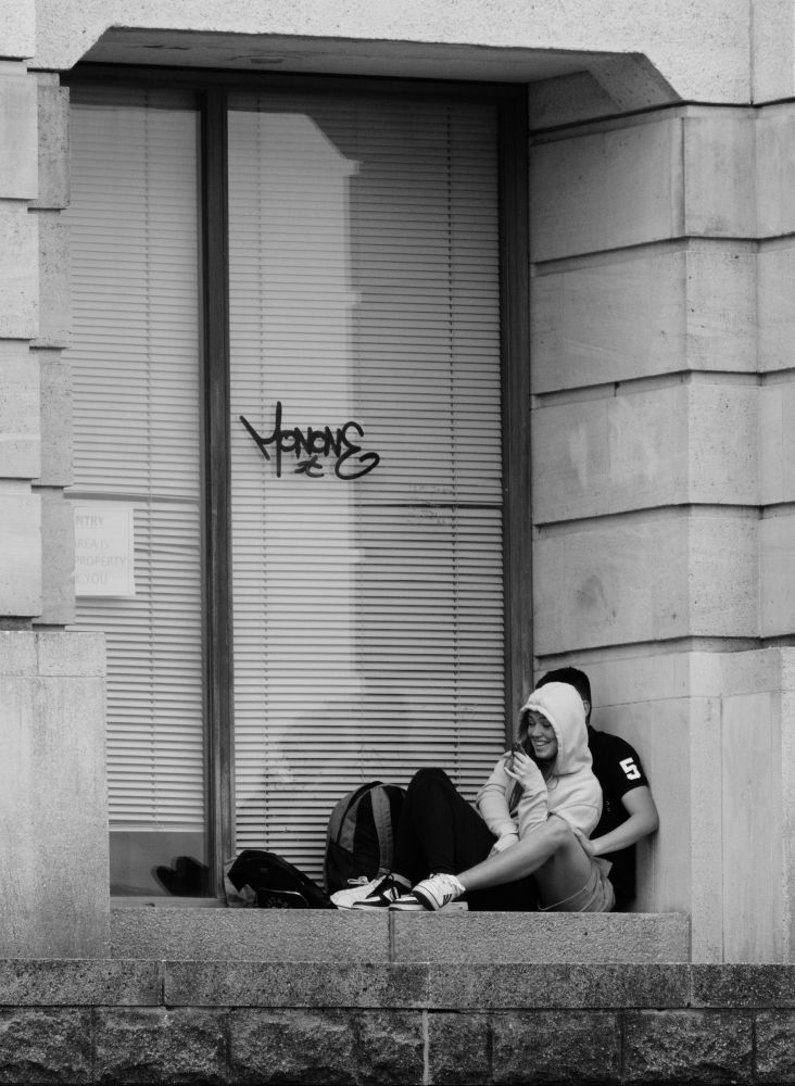 A black and white photography of a Boy and Girl sat in a window recess of the amphitheatre at Bristol Harbour. The young lady has a cookie in her hand and the Young man's face is hidden behind her.