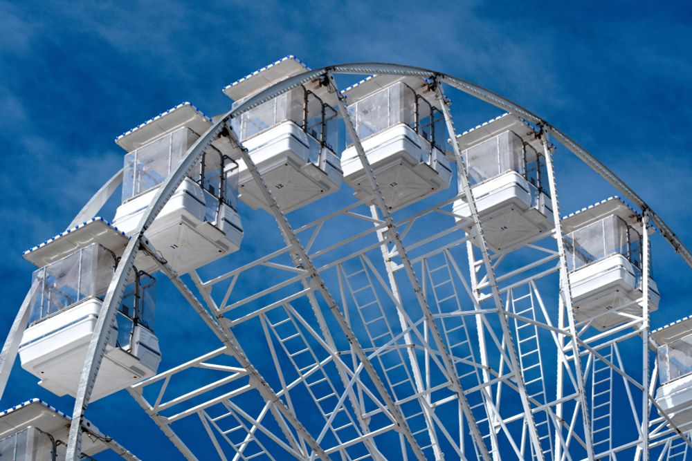 A colour photograph of a section of a white Ferris wheel. Pods 10 to 15 are in view at the top of the wheel arc. The sky is a rich blue with a wispy cloud layer. The sun is lowish and hitting from the left, illuminating the laddered frame work and pods.