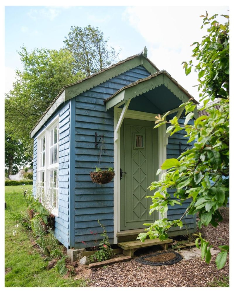 Exterior view of a light blue shed with a green door, situated in a garden setting. Hanging baskets and small plants add to the scene.