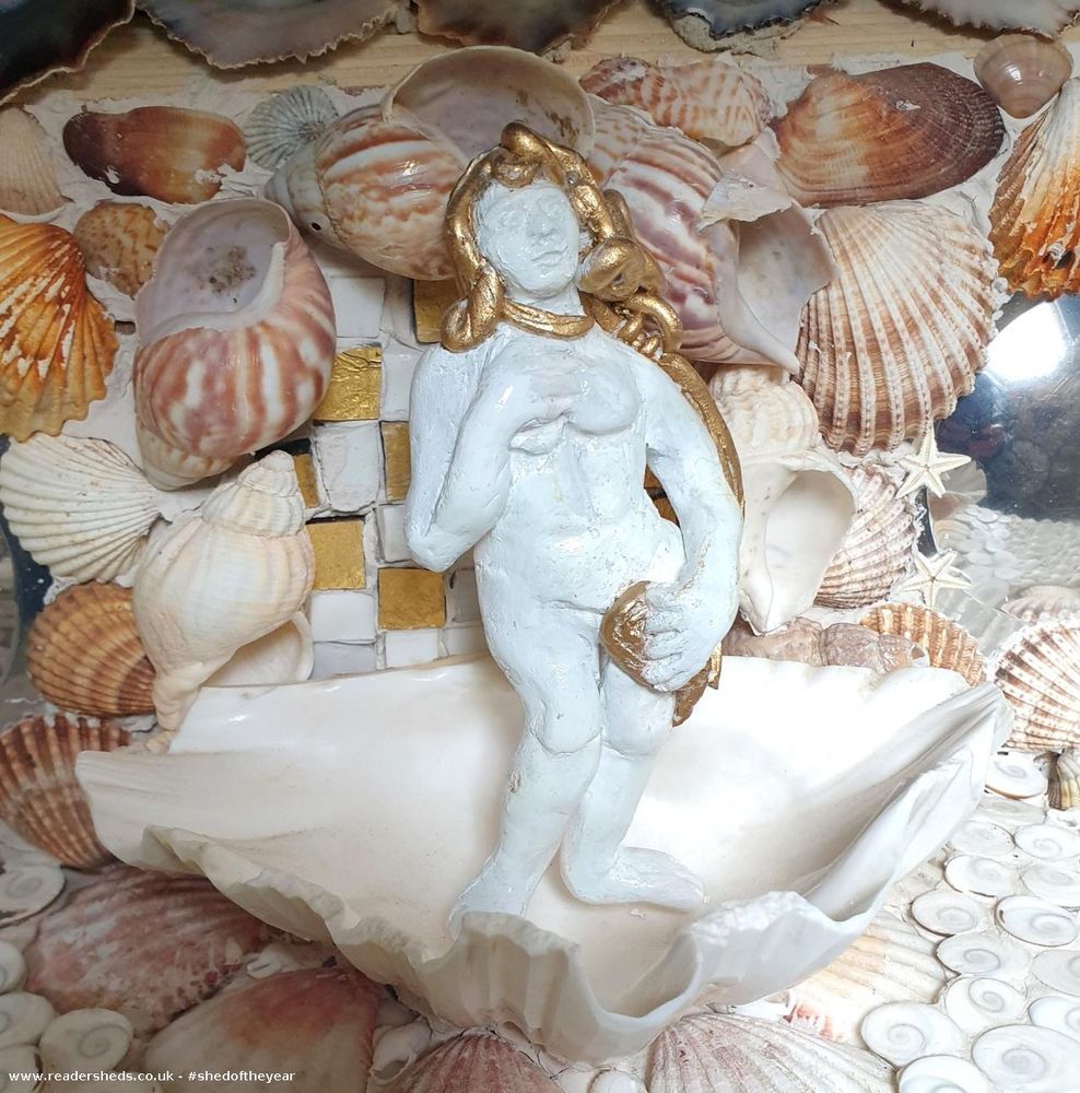 Close-up view of a decorative element within a grotto-like structure, featuring a plaster figure of a woman in a seashell, surrounded by various seashells, gold mosaic tiles, and gold accents. The figure appears to be inspired by the Venus archetype and represents a classical artistic representation.