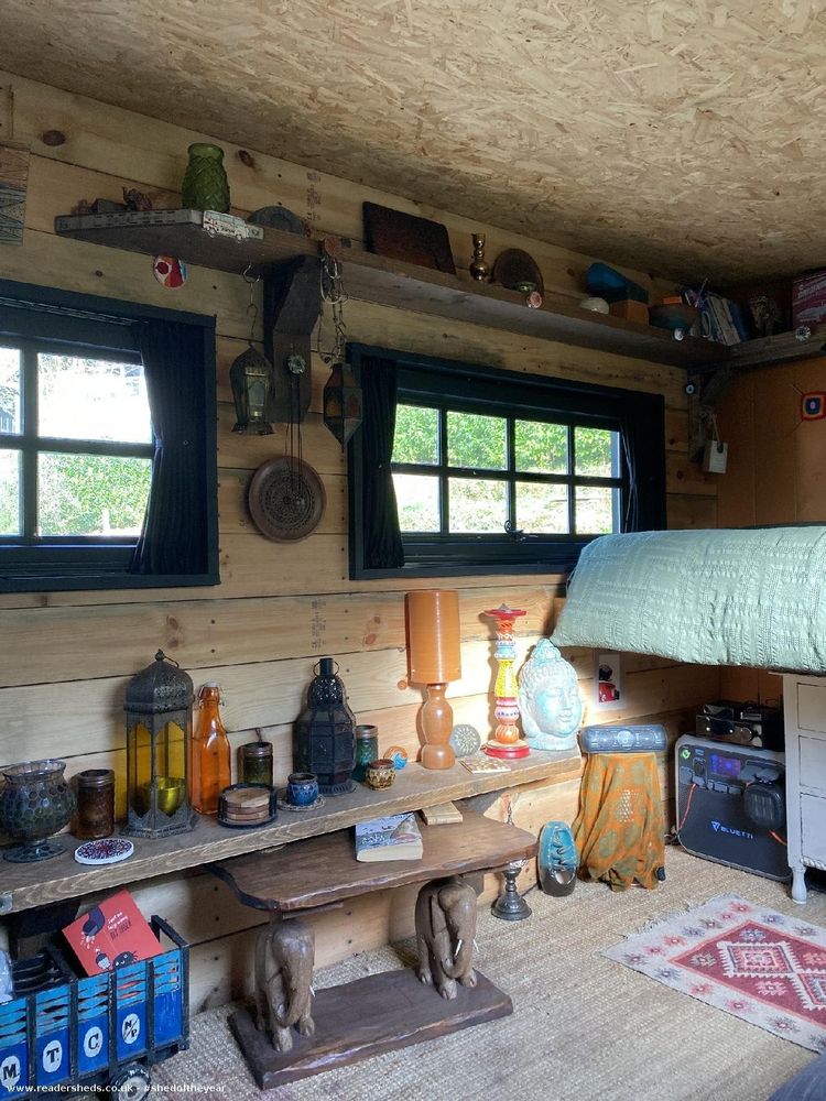 Inside a small cabin with wooden walls and OSB ceiling. A green-blanketed bed is elevated above a white dresser with colorful knobs. Below, a wooden shelf holds eclectic decor: colorful glass bottles, lanterns, a Buddha statue, and a lamp. A small table with books and elephant figurines sits on a woven rug with a patterned red mat. Two windows with black curtains allow natural light to enter.