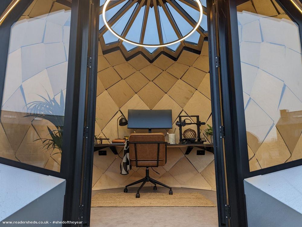 The interior of a finished geodesic dome, set up as a home office. A desk with a computer, chair, and 3D printer is centered in the room. The walls are finished with light wood panels in a geometric pattern. Black-framed glass doors are open to the outside.