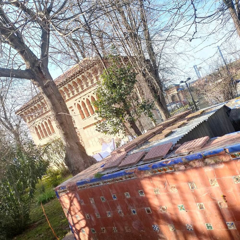 Toledo, Spain - Partially obscured view of a brick building featuring arched windows framed by bare trees. Decorated red stone building and pool structures with decorative colorful tiles on the top of each other. A corner of bridge can be seen from in the trees behind as well.

Photo 3: