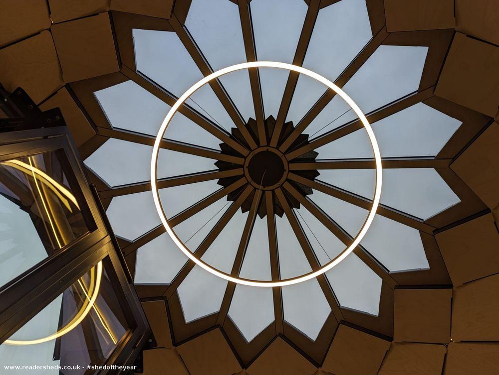 Looking up at the completed interior of a geodesic dome. A large circular light fixture dominates the ceiling, which is made of triangular wooden panels. A portion of a black-framed glass elevator or enclosure is visible in the lower left corner.