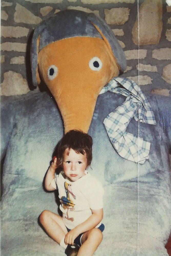 A young child is sitting on a large, plush blue couch in front of a giant stuffed animal that resembles an anteater or aardvark with a long orange snout and big round eyes but is in fact a Womble, The child is wearing a white T-shirt with a cartoon bird on it (Big Bird, I loved that tshirt) and blue shorts.  The background features a wall with a stone pattern.
