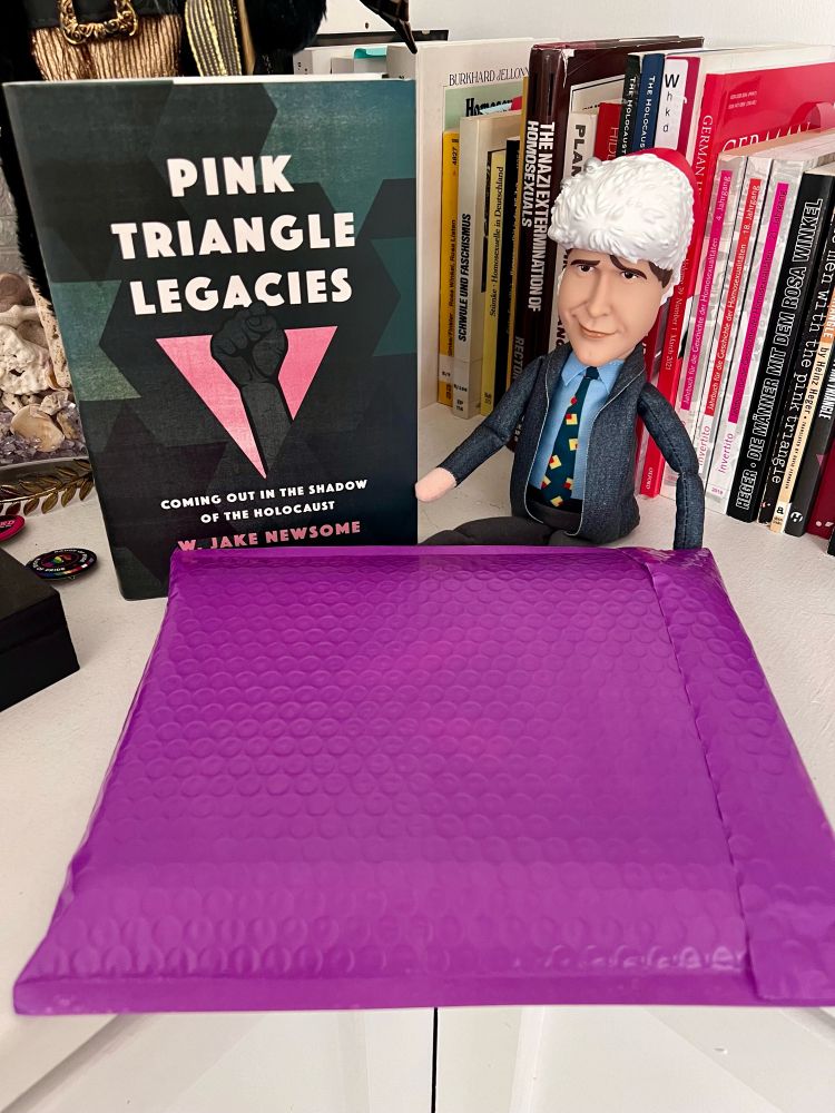 A purple bubble mailing envelope sits in front of a book with the title “Pink Triangle Legacies: coming out in the shadow of the Holocaust.” Next to the book is a small doll of the character Clark Griswald from the movie “Christmas Vacation”