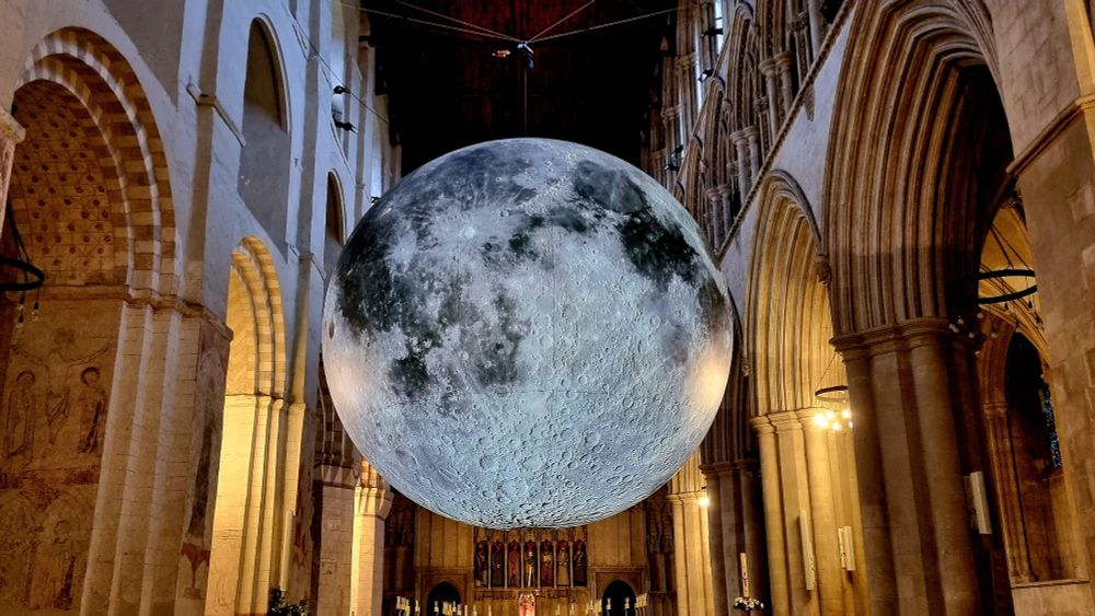 Art installation. 7m diameter sphere depicting the surface of the moon with craters and lakes suspended from the ceiling in St Albans cathedral. The moon looks like it almost touches the arches of the cathedral's nave.