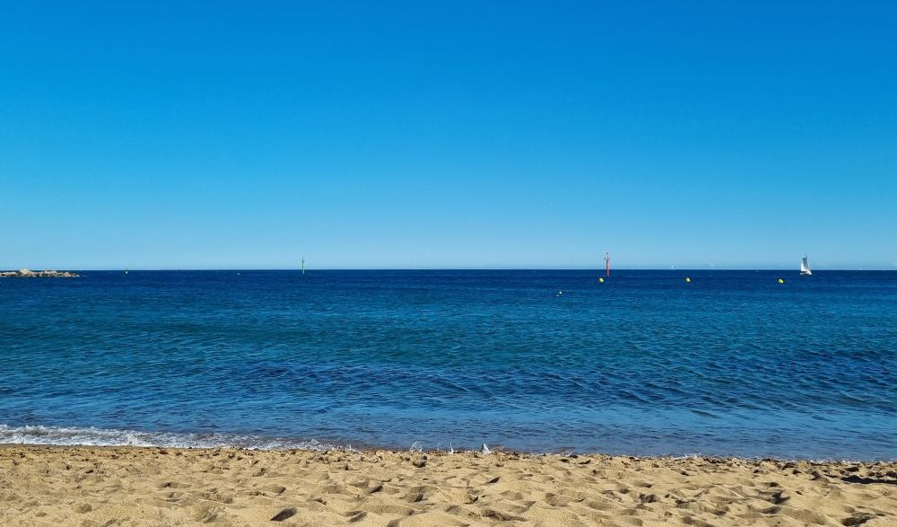 Panorama picture of the blue slightly wavy Mediterranean Sea enclosed by light brown sand at the bottom and a cloudless blue sky above
