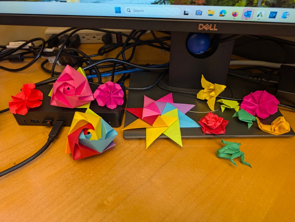 A desk, lit oh so flourescently, and the base of a computer monitor that is littered with origami projects: flowers, critters, and a huge rainbow star