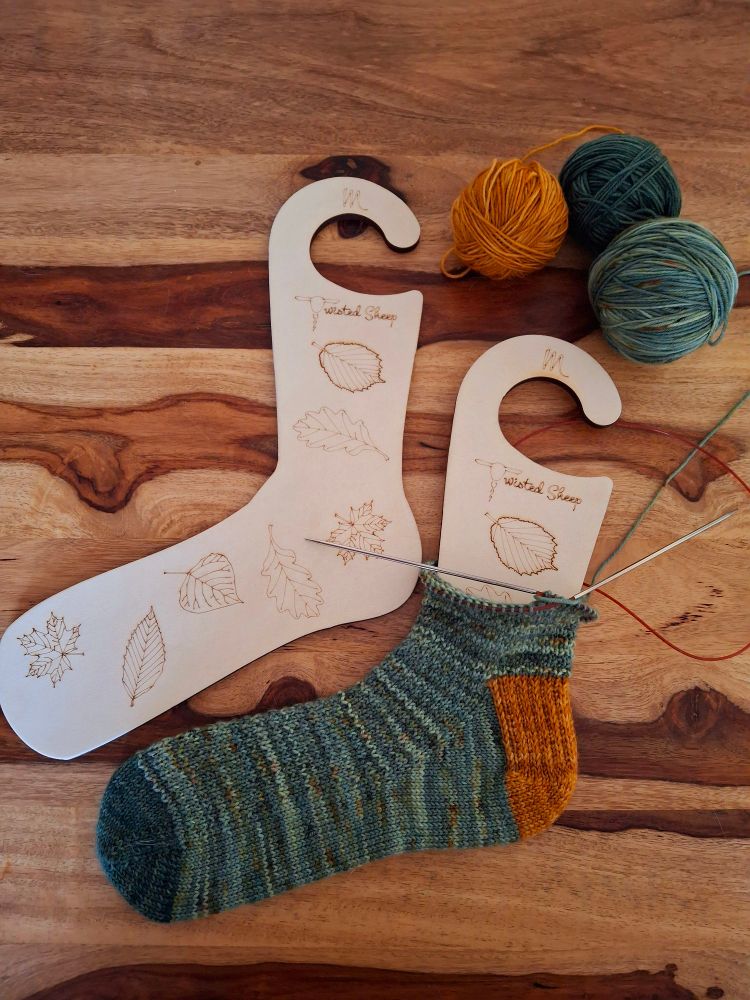 A set of sock blockers on a wooden table. The blockers are made of wood and decorated with line drawings of leaves. On the right sock blockers is a green sock in progress. The sock has a mustard coloured heel flap. A ball of mustard yarn and two balls of green yarn are at the top right of the image.