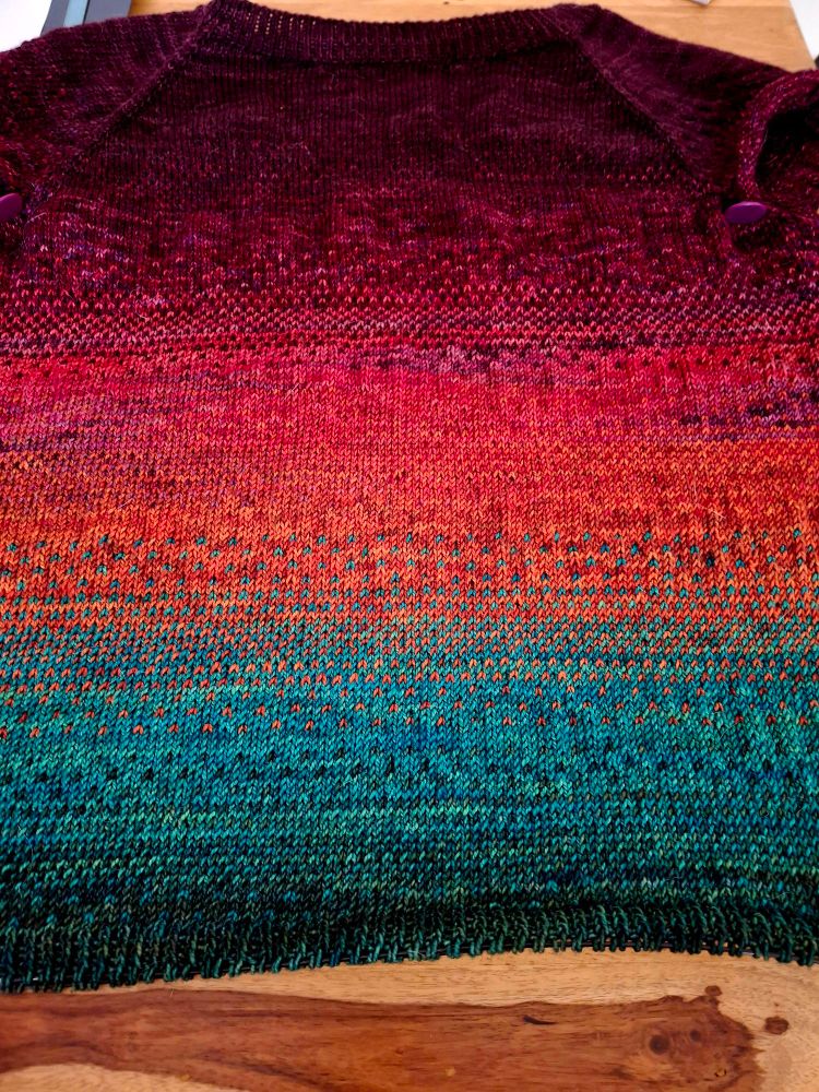 A hand knitted sweater in progress. It is knit from the top down and features bands of colour knit in a fade stitch pattern. It starts with purple and moves through dark pink, light pink, orange, bright green, and dark green.