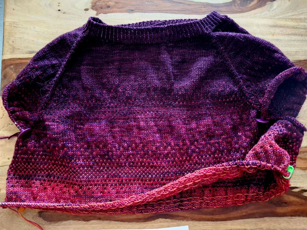 A hand knitted raglan sweater in progress. It is knit from the top down. The colour transitions from purple at the top, through to pink and then orange.