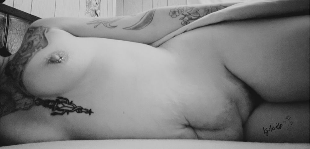 An alternative MILF is lying on her side on the bed, nude exposing various tattoos, small pierced breasts, mom belly and a little bit of bush. 