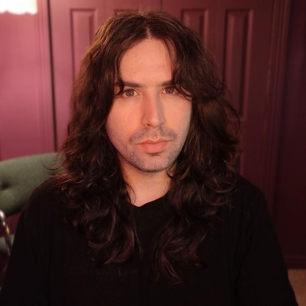 Stephen, a white-japanese nonbinary person, sitting at their desk, getting ready to go live on twitch. stephen has long curly brown hair and is wearing all black.
