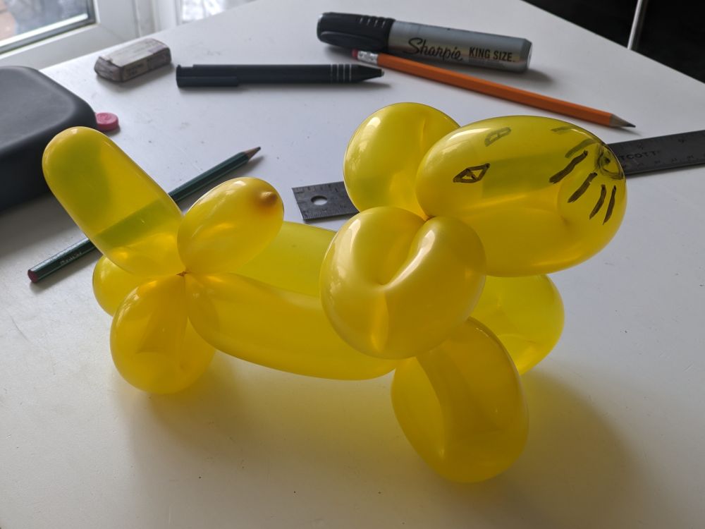 A yellow balloon animal (poorly?) shaped like a cat. There are whiskers and squinting eyes drawn with sharpie where the face would be.