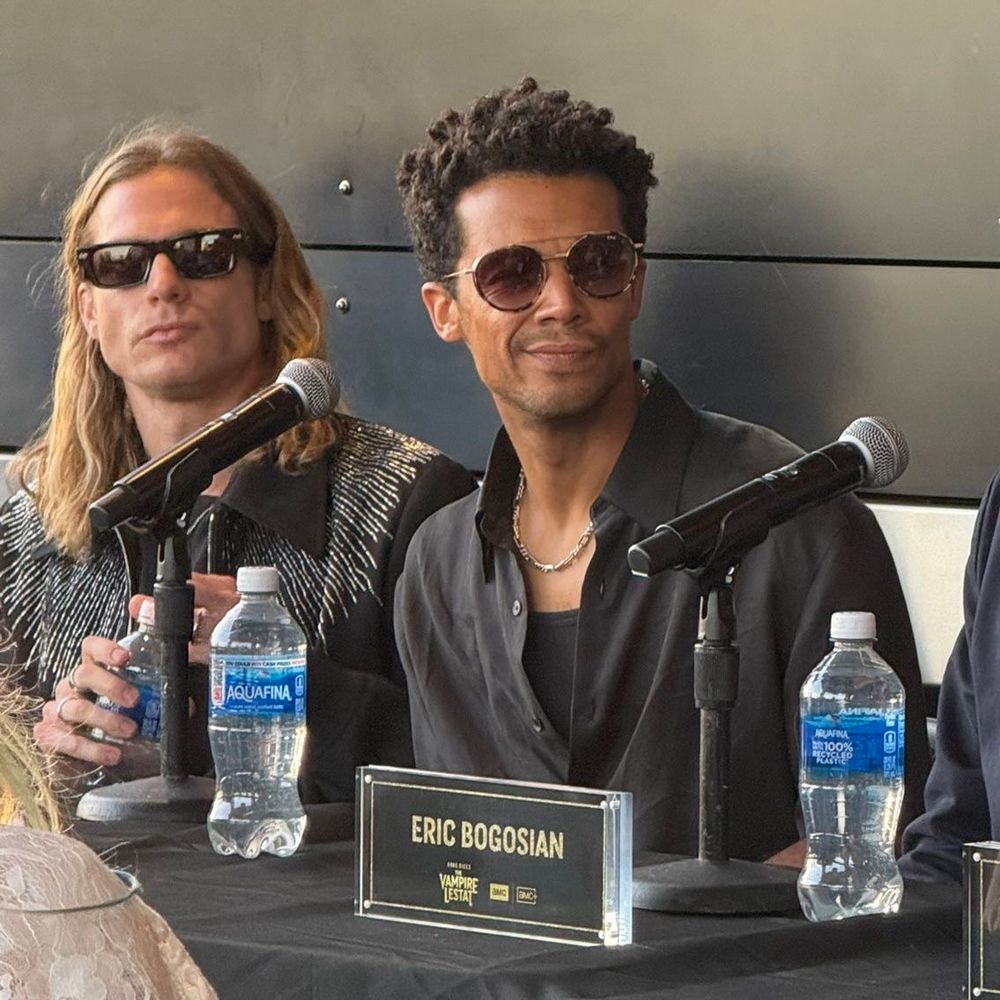 on the left sam reid sits behind a table with a microphone on it, in front of him. he holds a water bottle and is wearing sunglasses. next to him sits jacob anderson wearing sunglasses too, who also has a microphone in front of him and a water bottle next to it. 